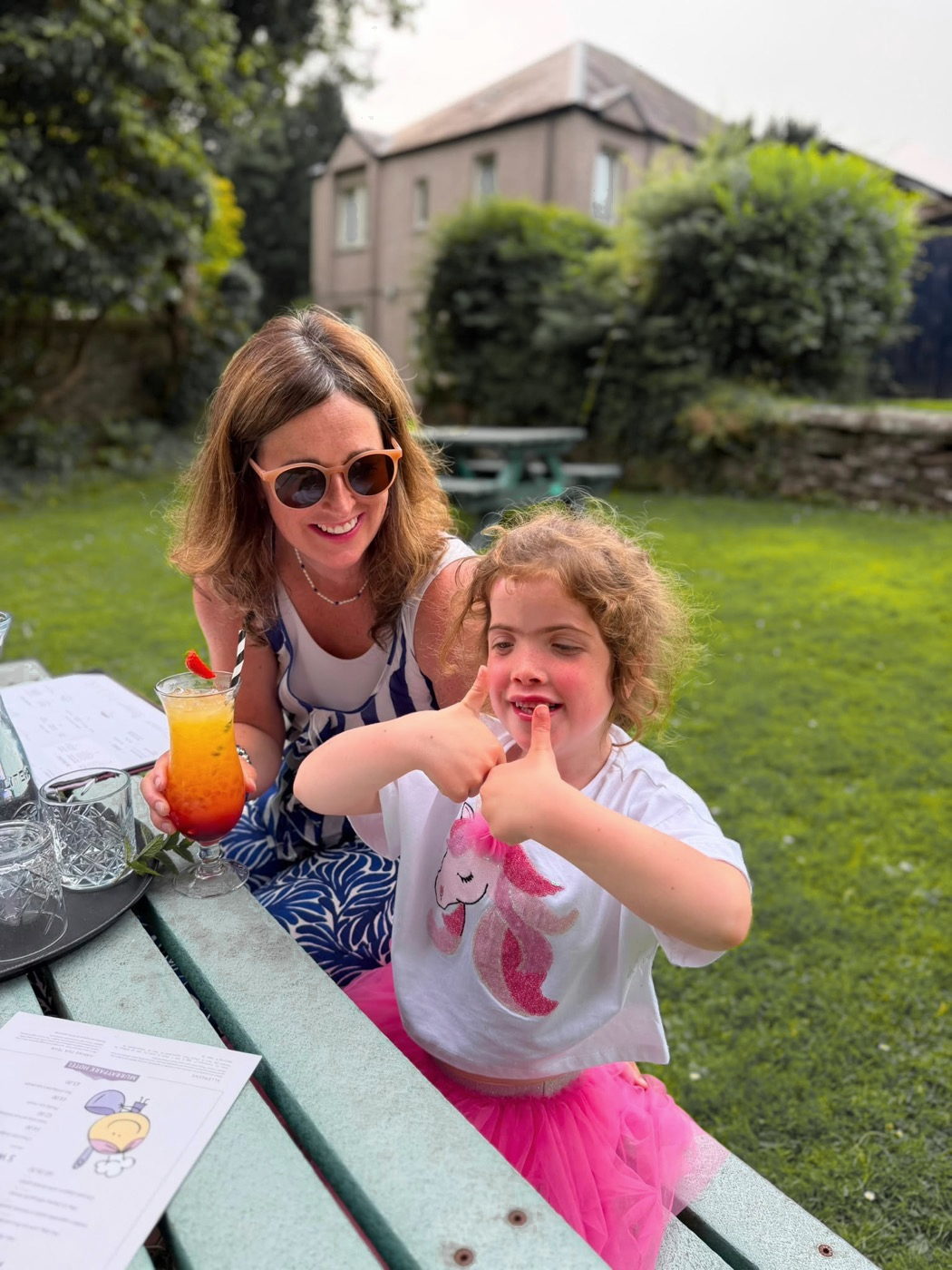 Aoife signing with both thumbs up, sitting with her mum Holly at an outdoor table.
